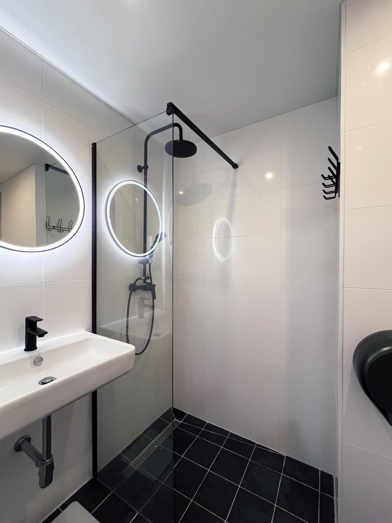 Bathroom in Solospace Gym featuring a modern design with a walk-in shower, a rain showerhead, a glass partition, a circular backlit mirror above a rectangular sink with a black faucet, and dark tiled flooring