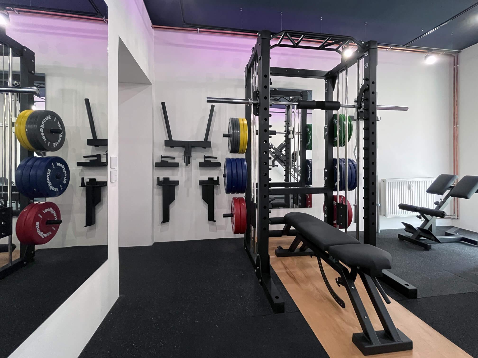 Interior of Solospace Gym featuring a squat rack with an integrated Smith machine, attachments mounted on the wall, an adjustable incline bench with a foot catch for decline movements, a deadlift platform, and a hyperextension bench in a well-lit room with rubber flooring