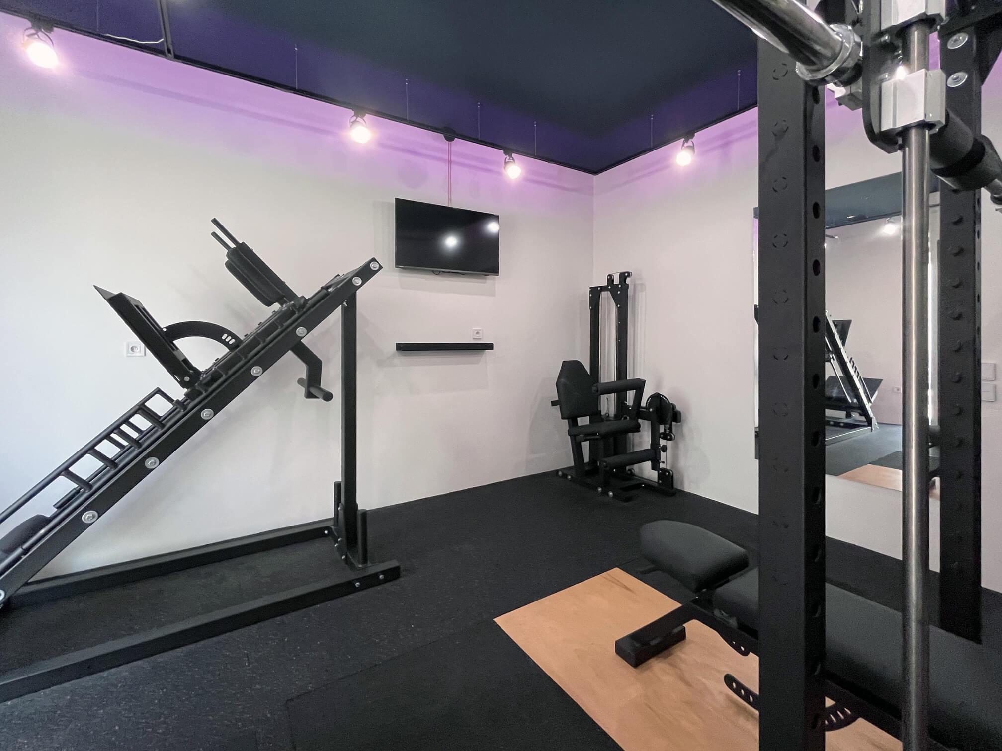 Interior of Solospace Gym featuring a Leg Press and Hack Squat combo machine on the left, a seated Leg Curl and Extension combo machine on the right, and in the foreground, a Squat Rack with an integrated Smith machine, an adjustable incline bench with a foot catch for decline movements, and a deadlift platform. The room is well-lit with rubber flooring and overhead lights.