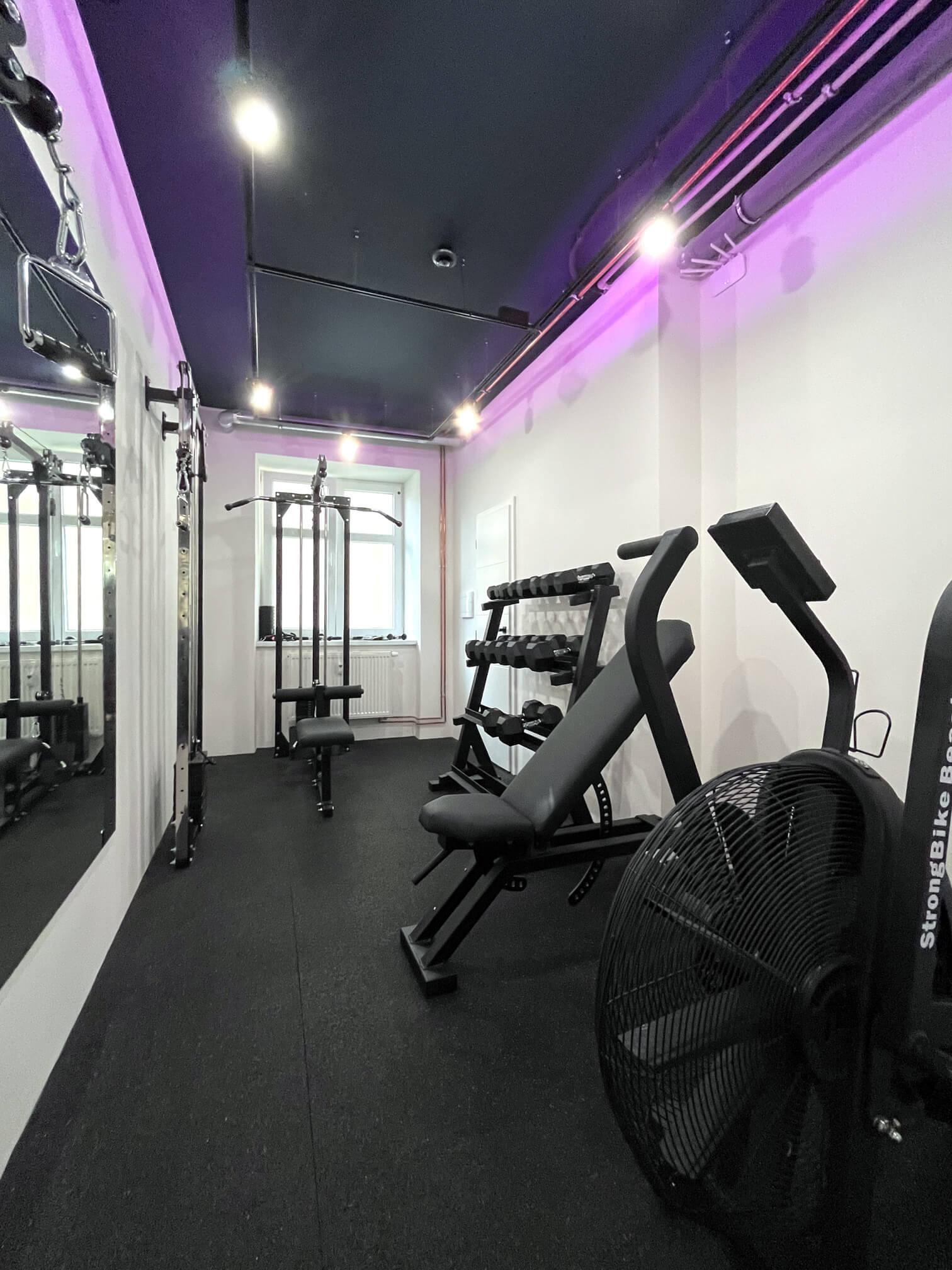 Interior of Solospace Gym featuring an airbike, an adjustable incline bench, a set of dumbbells on a rack, a Lat pulldown and seated row machine in the background, and a cable crossover machine next to the mirror. The room is well-lit with overhead lighting and has rubber flooring