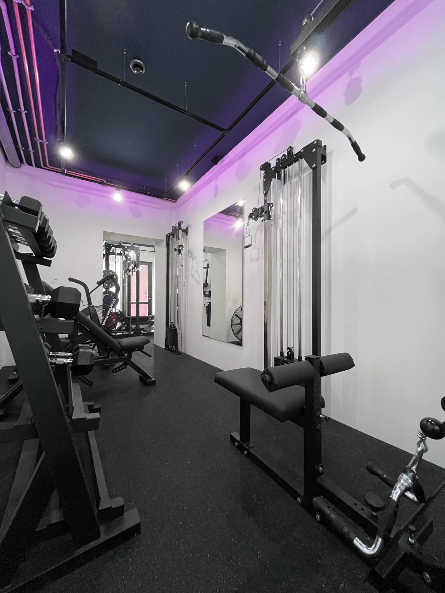 Interior of Solospace Gym featuring a cable crossover machine, an adjustable incline bench, dumbbells on a rack, an airbike, a Lat pulldown and seated row machine, and in the far background, a Squat Rack with an integrated Smith machine. The room is well-lit with overhead lights, rubber flooring, and a large mirror reflecting parts of the gym equipment